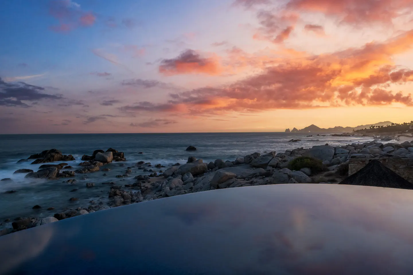 Infinity pool overlooking a rocky coastline at sunset with a vibrant orange and purple sky over the ocean.