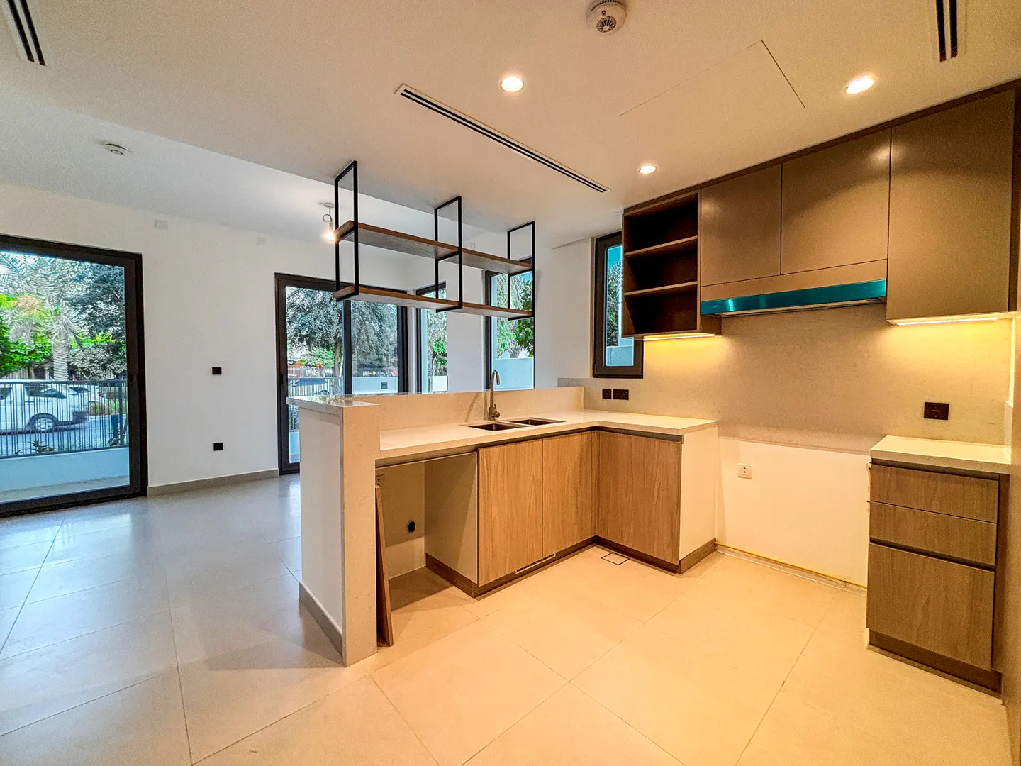 A modern kitchen with light wood cabinets, white countertops, and black metal shelving. Large sliding glass doors lead to an outdoor area.
