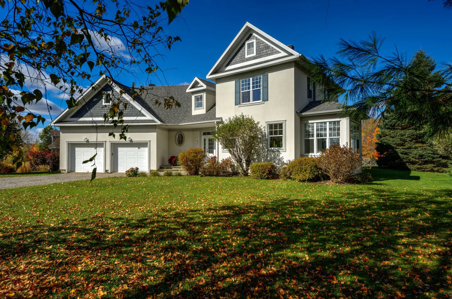Two-story house with a two-car garage, gray roof, and white exterior. The lawn is green with fallen leaves.