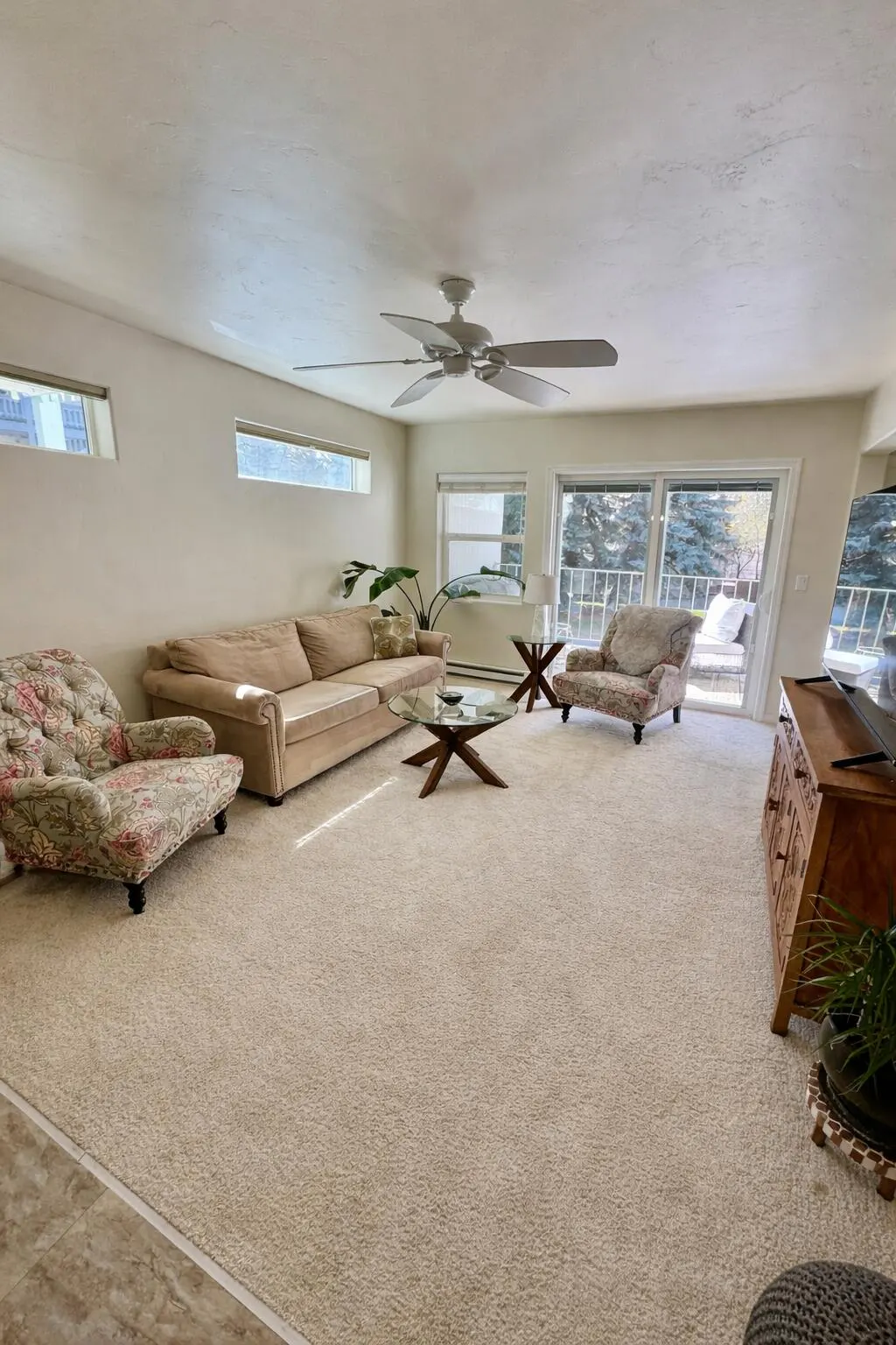 Living room with beige carpet, sofa, floral armchairs, glass coffee table, and a ceiling fan. Sliding glass doors lead to a balcony.