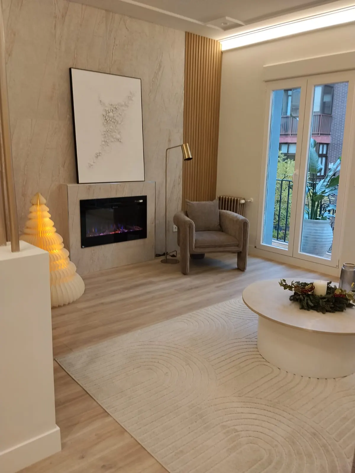 A modern living room with a fireplace, art, and a beige armchair. A round coffee table sits on a patterned rug. Natural light streams through the windows.