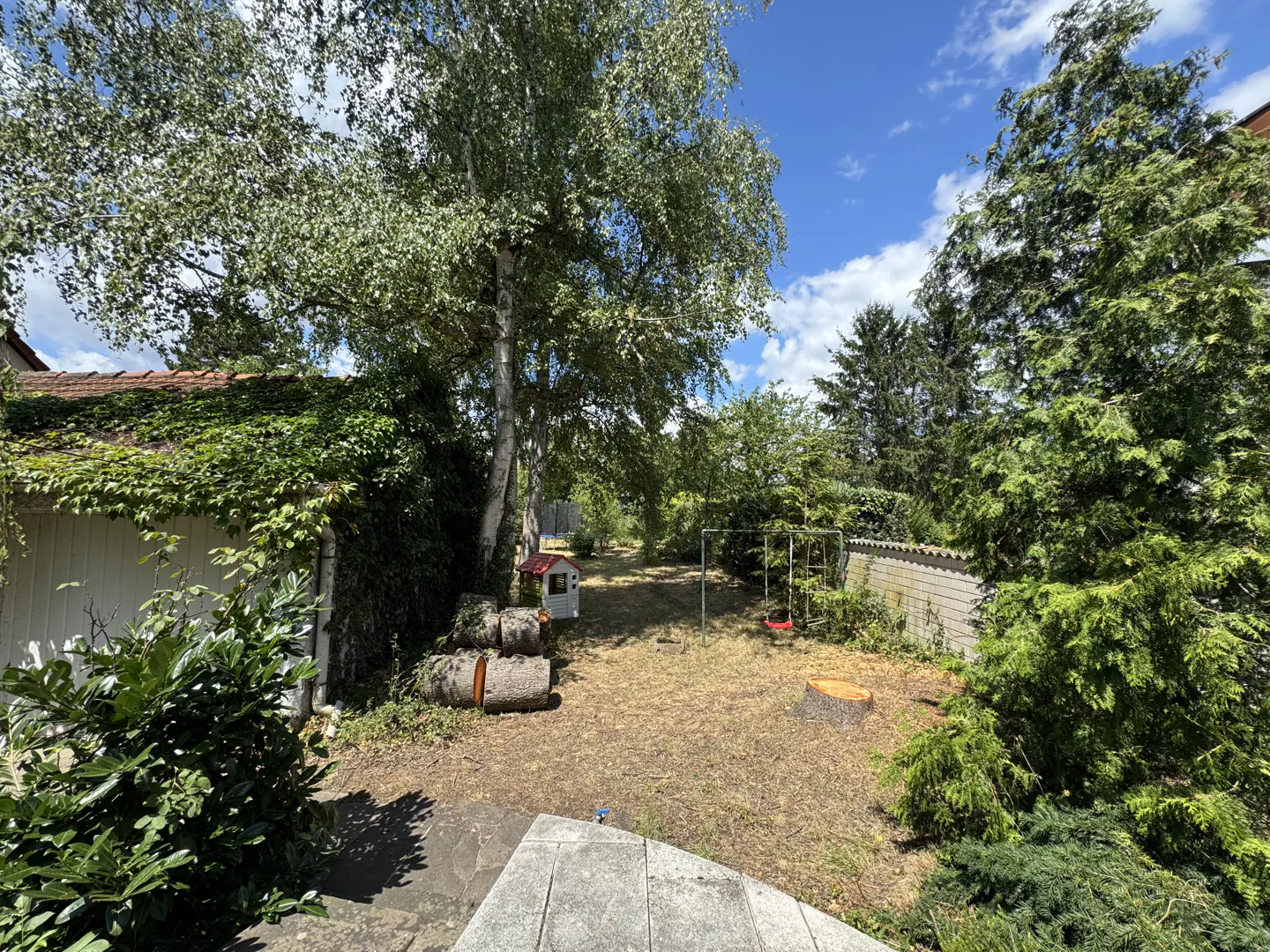 A backyard with trees, a swing set, a playhouse, and stacked logs on a sunny day.