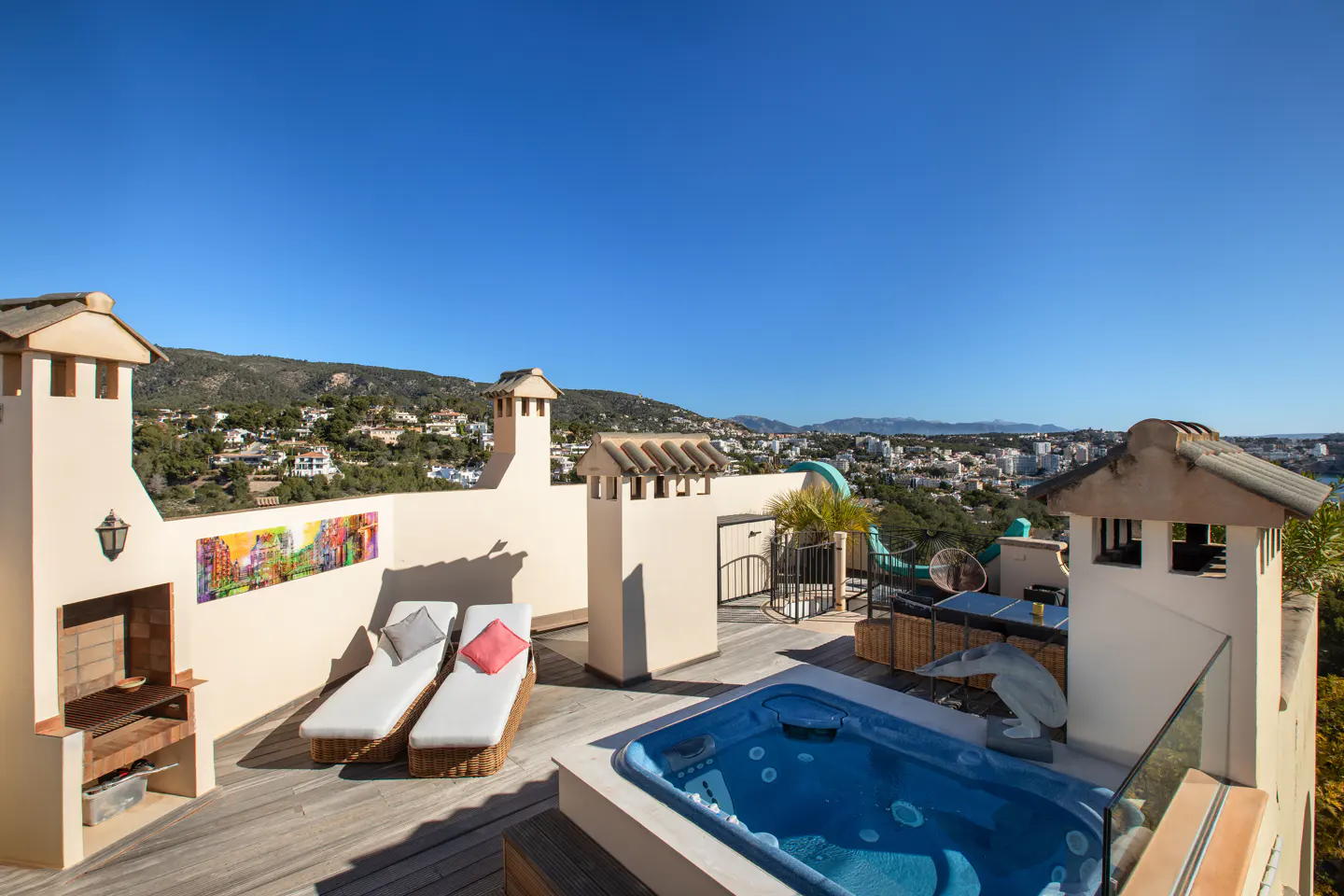 Rooftop patio with a blue hot tub, two lounge chairs, and a grill. A colorful painting hangs on the wall. City and mountain views in the background.