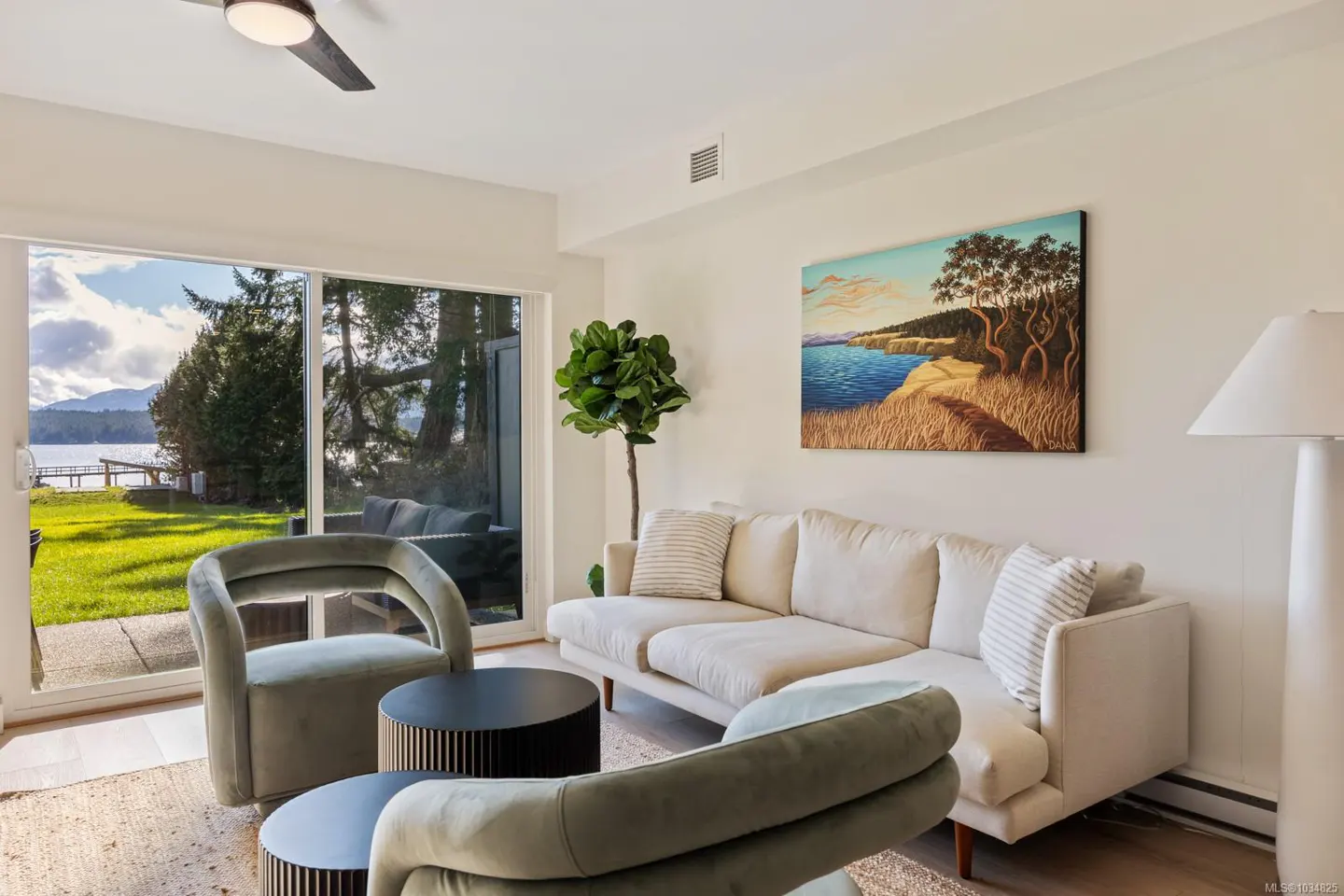 Bright living room with a cream sofa, two green chairs, and a painting of a coastal scene. A large window shows a grassy lawn and a lake.