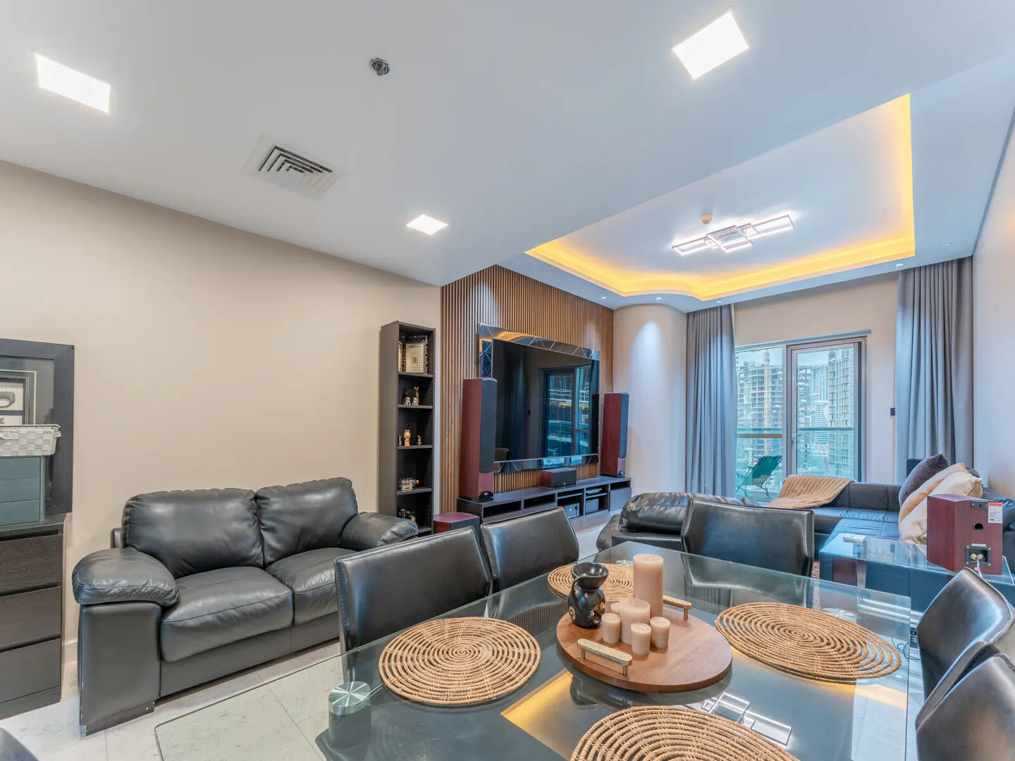 Living room with black leather sofas, glass dining table, TV, and city view through the window.