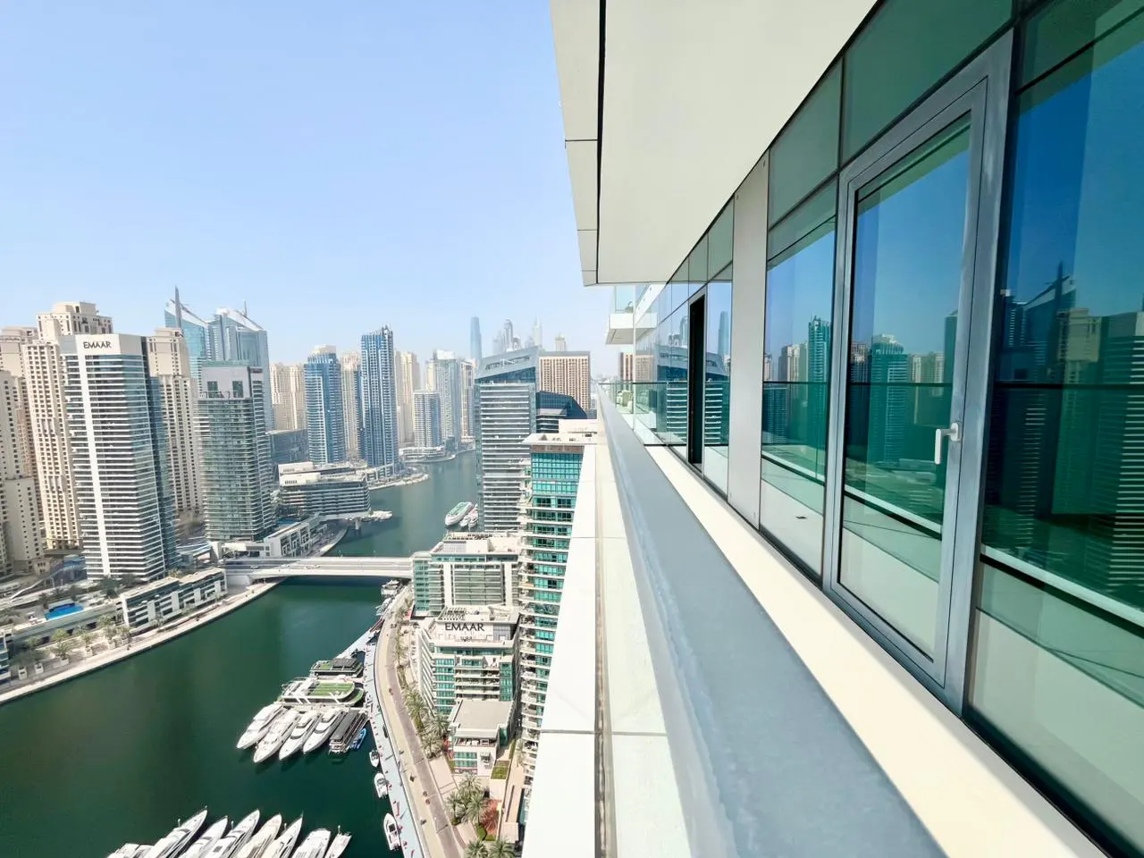 View from a high-rise balcony overlooking Dubai Marina, with skyscrapers, water, and yachts visible.