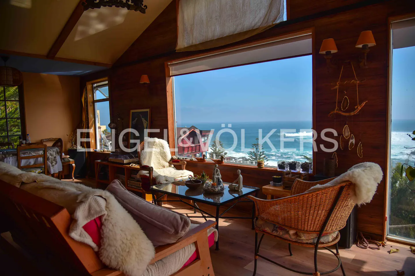 Living room with wood walls, a glass coffee table, and a large window overlooking the ocean. Wicker chairs and a wooden sofa are present.