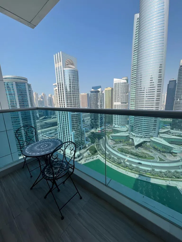 Balcony view of Dubai skyline. Black metal table and chairs sit on a gray wood balcony overlooking skyscrapers and green waterways.