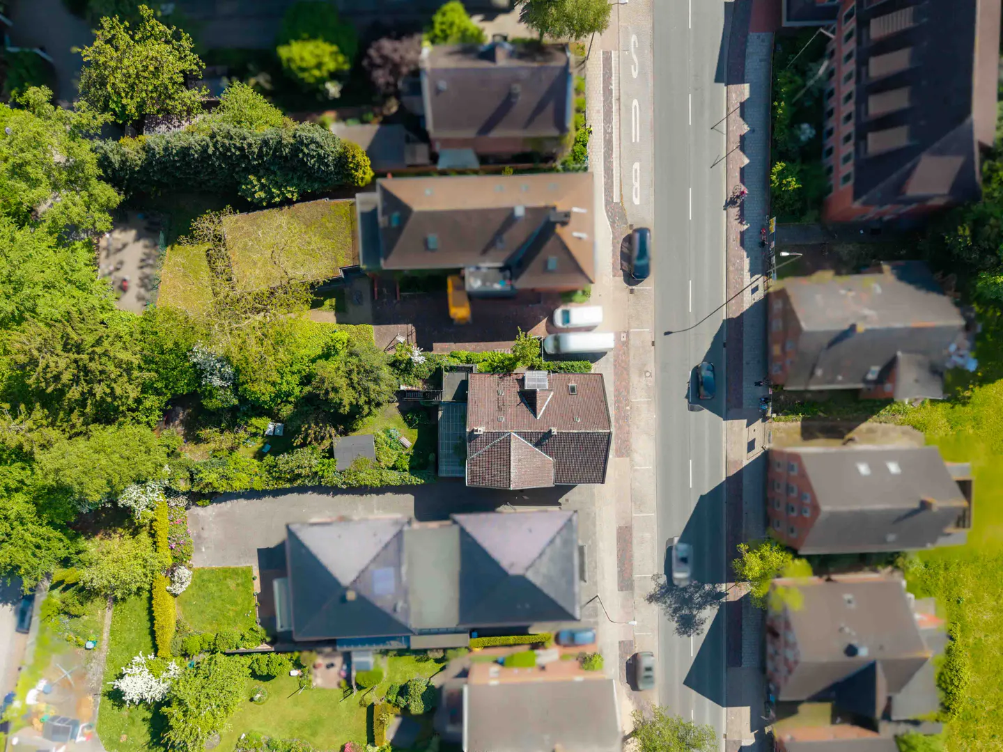 Aerial view of a residential street with houses, green trees, and cars parked along the road.