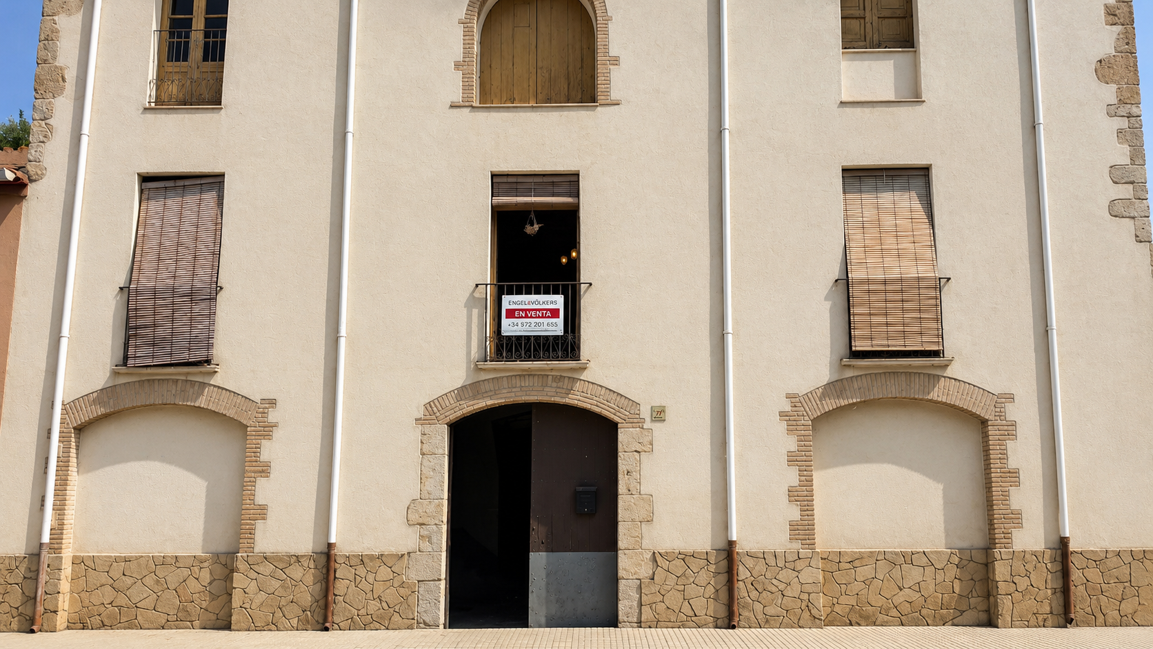 Cream-colored stone building with a central entrance, arched windows, and a "For Sale" sign on a small balcony. [AI-generated text] [Modificat per IA]