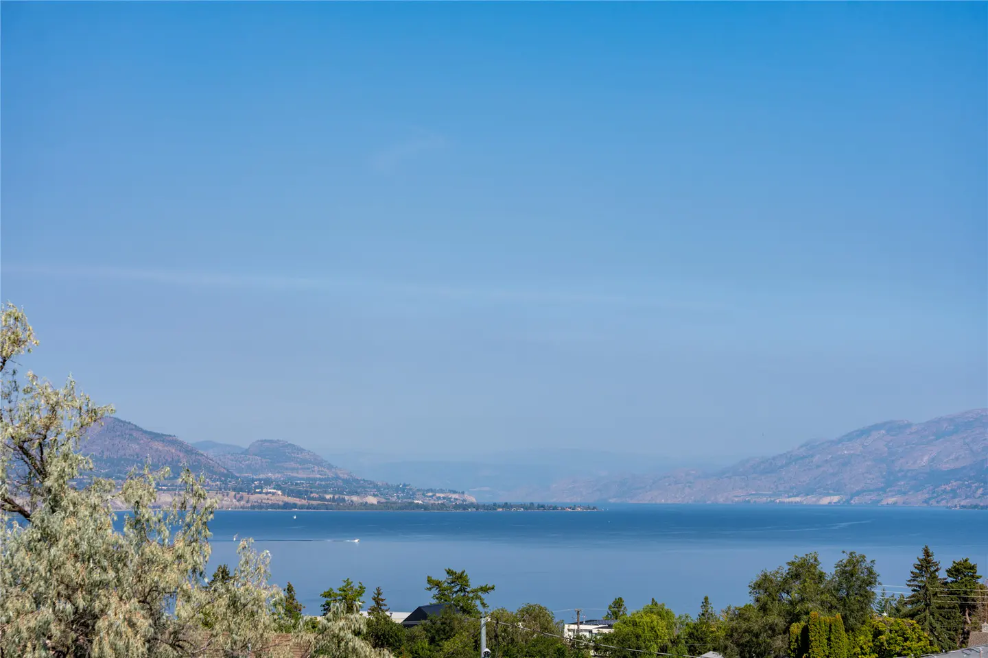 Scenic view of a blue lake surrounded by mountains under a clear blue sky. Green trees are visible in the foreground.