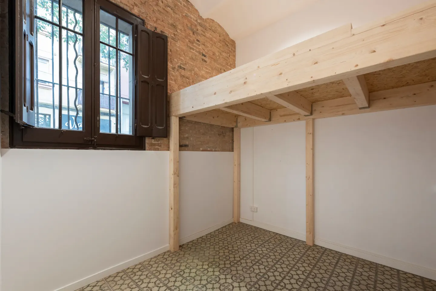 A bright room with a loft bed frame, white walls, and patterned tile floor. A window with brown shutters is on the left, and exposed brick is visible.