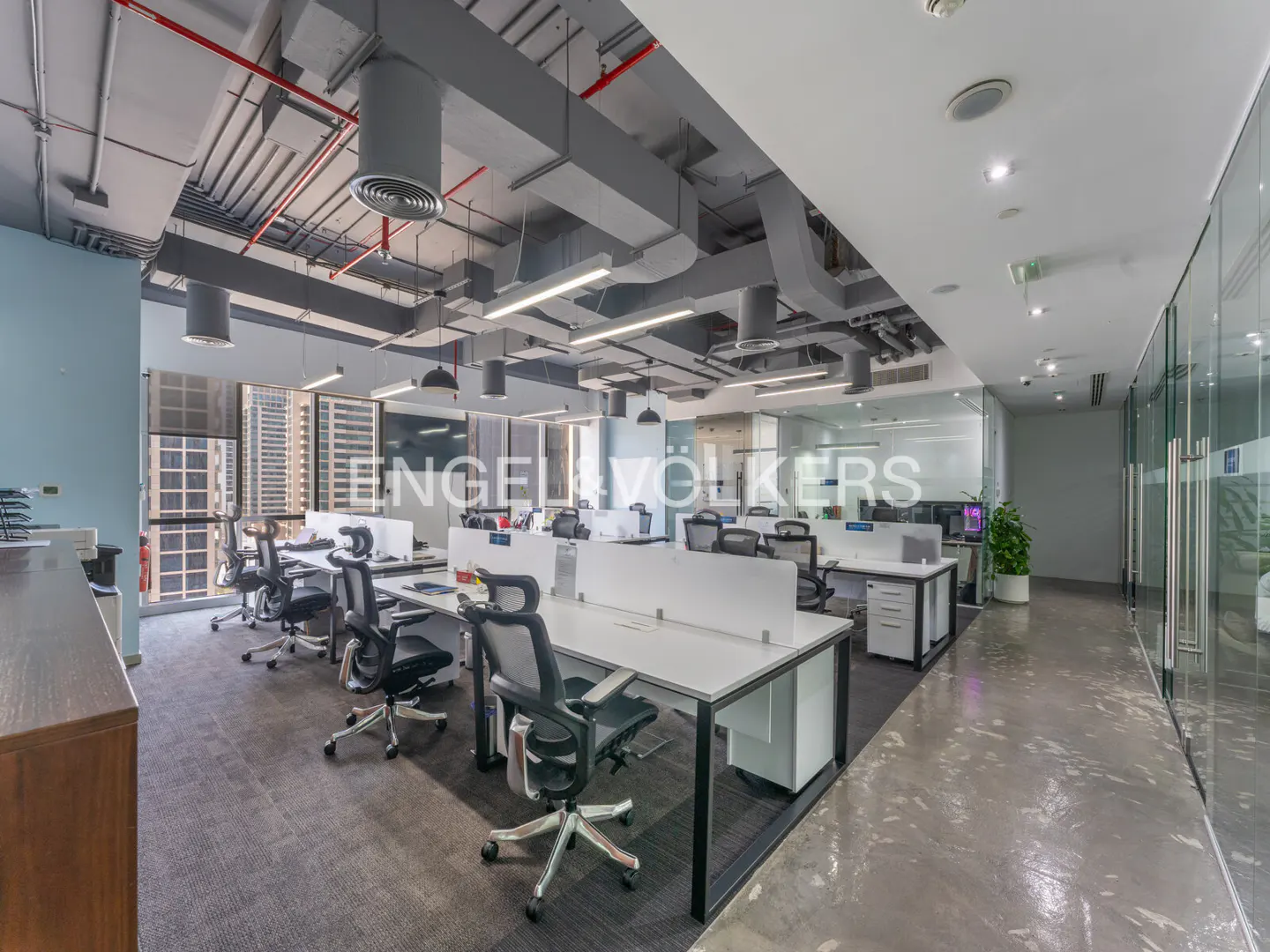 Bright, modern office space with white desks, black chairs, and gray carpet. Exposed ceiling with ductwork and lighting. Glass-walled offices visible.