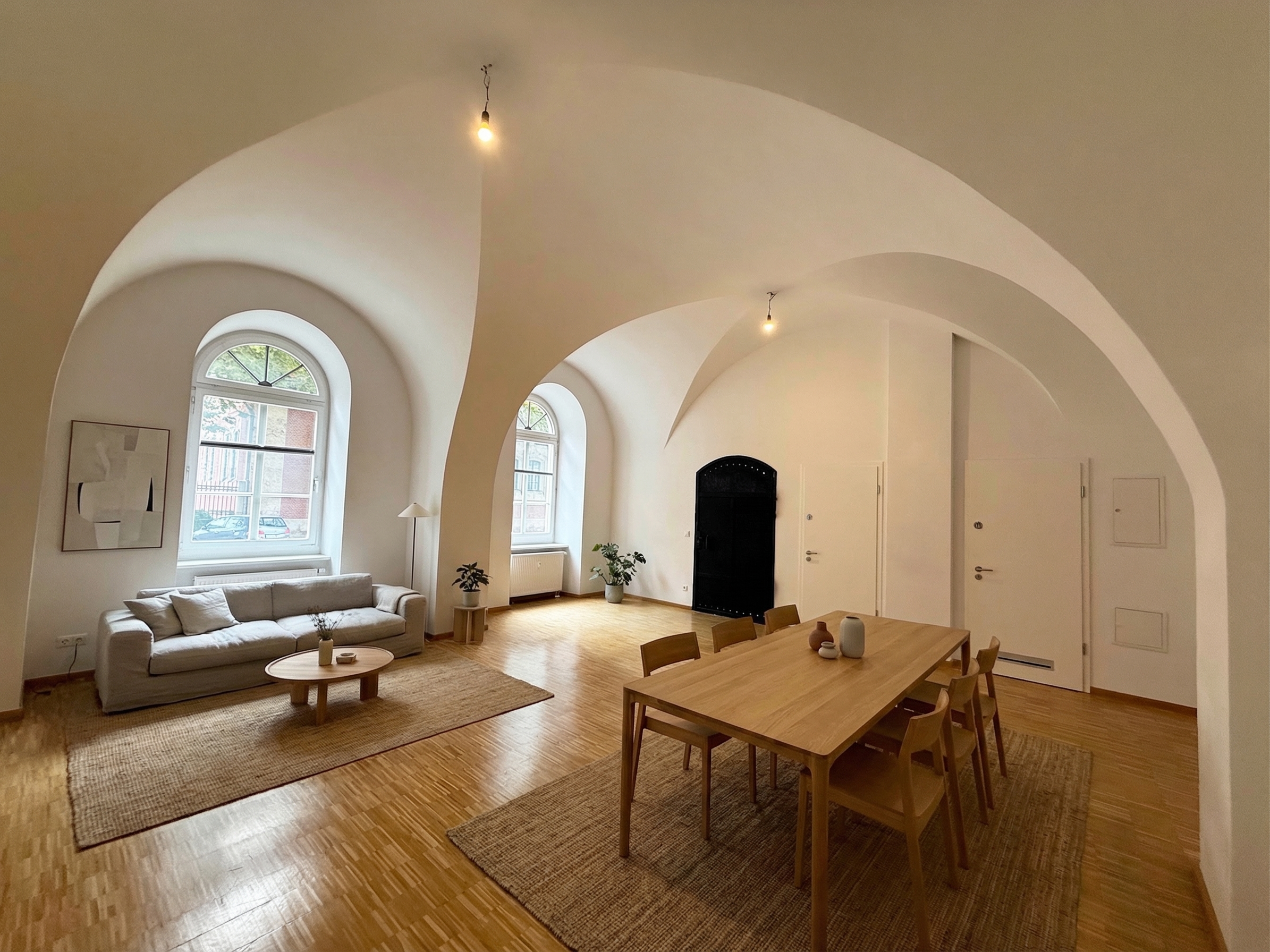 Bright, airy living space with arched ceilings, wood floors, and jute rugs. A sofa, table, and dining set furnish the room. Large windows let in natural light.
