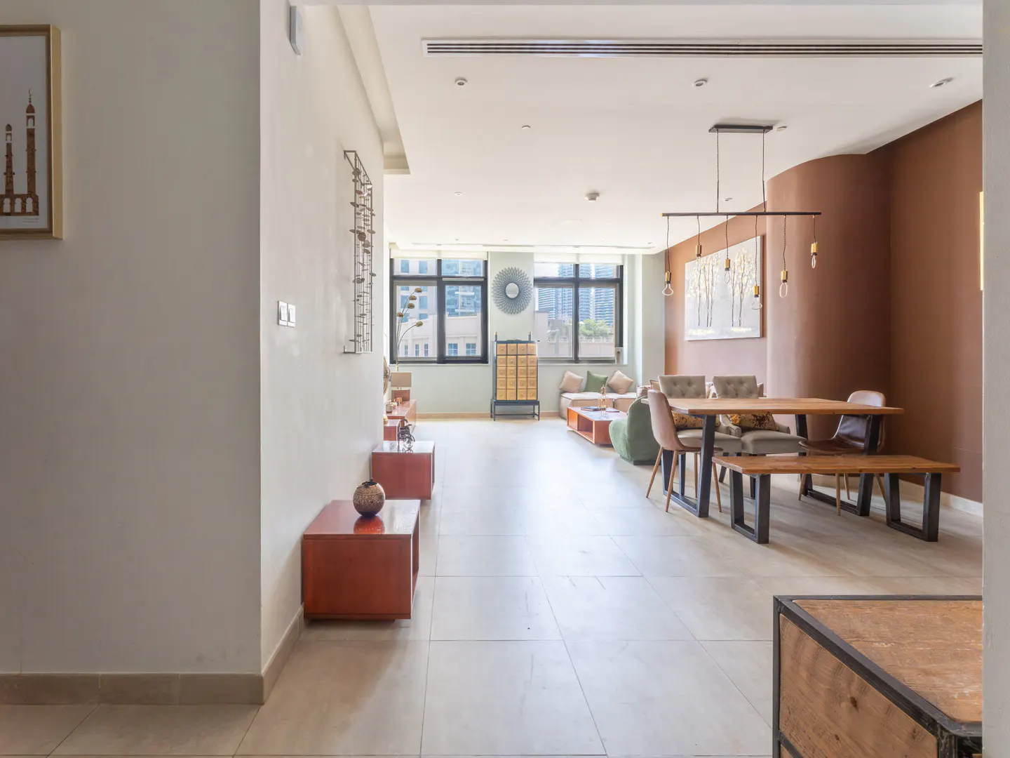 A modern apartment interior with a dining table, bench, chairs, and living area with a sofa and windows.