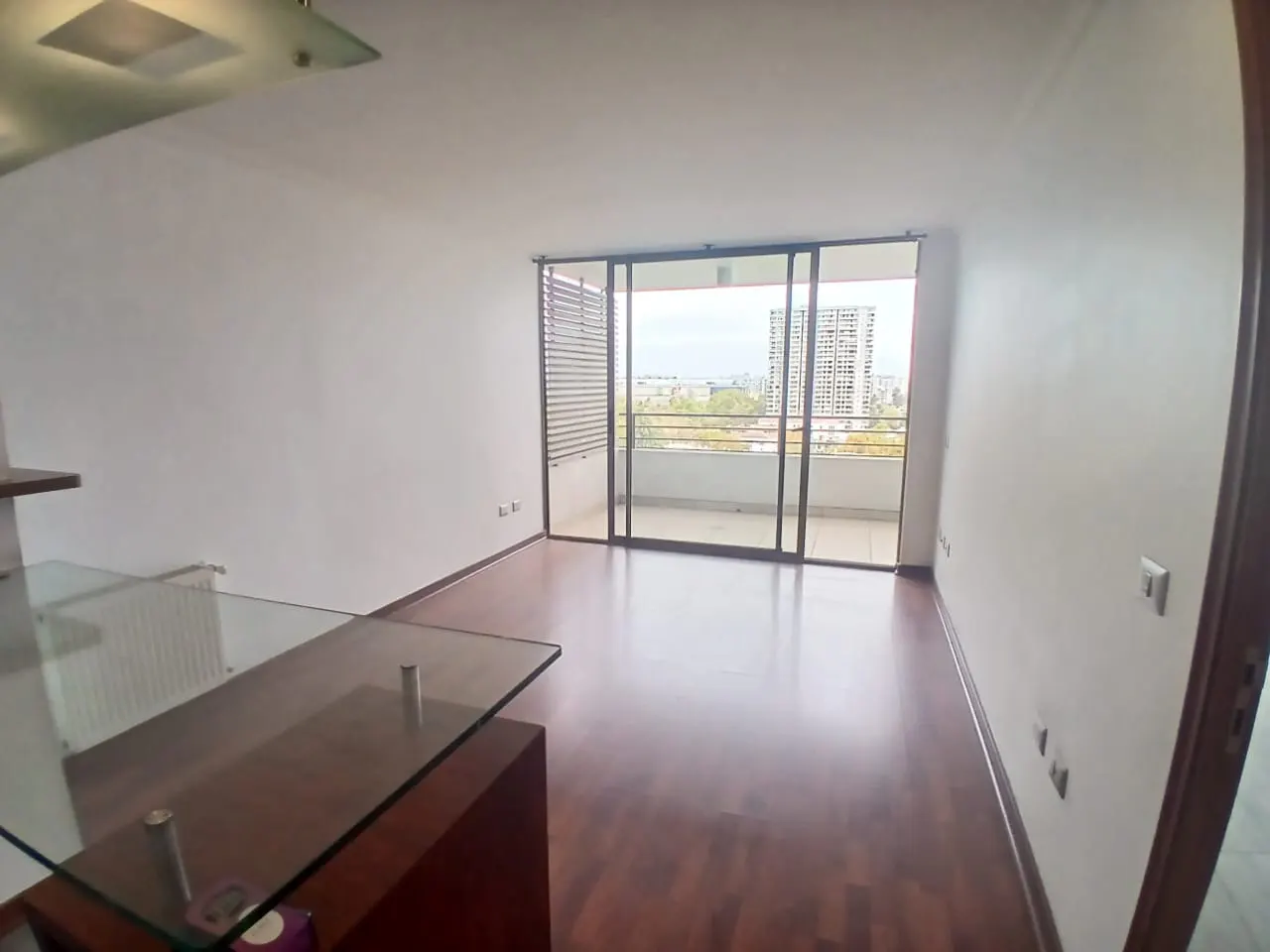 A bright, empty living room with hardwood floors and white walls. Sliding glass doors lead to a balcony with a city view.