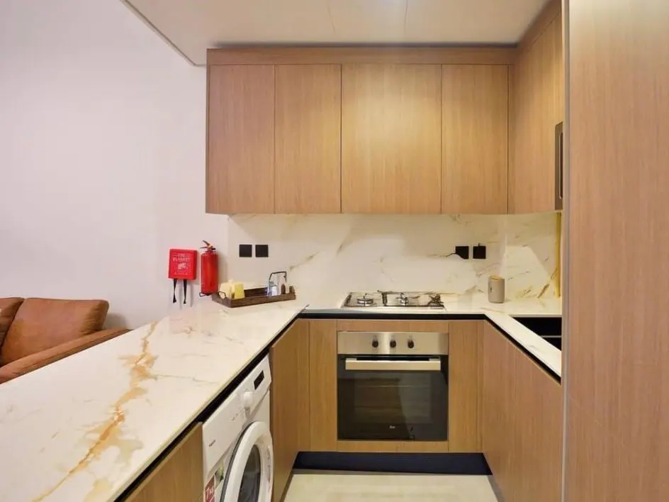 A modern kitchen with light wood cabinets, marble countertops, and stainless steel appliances. A washing machine is visible on the left.