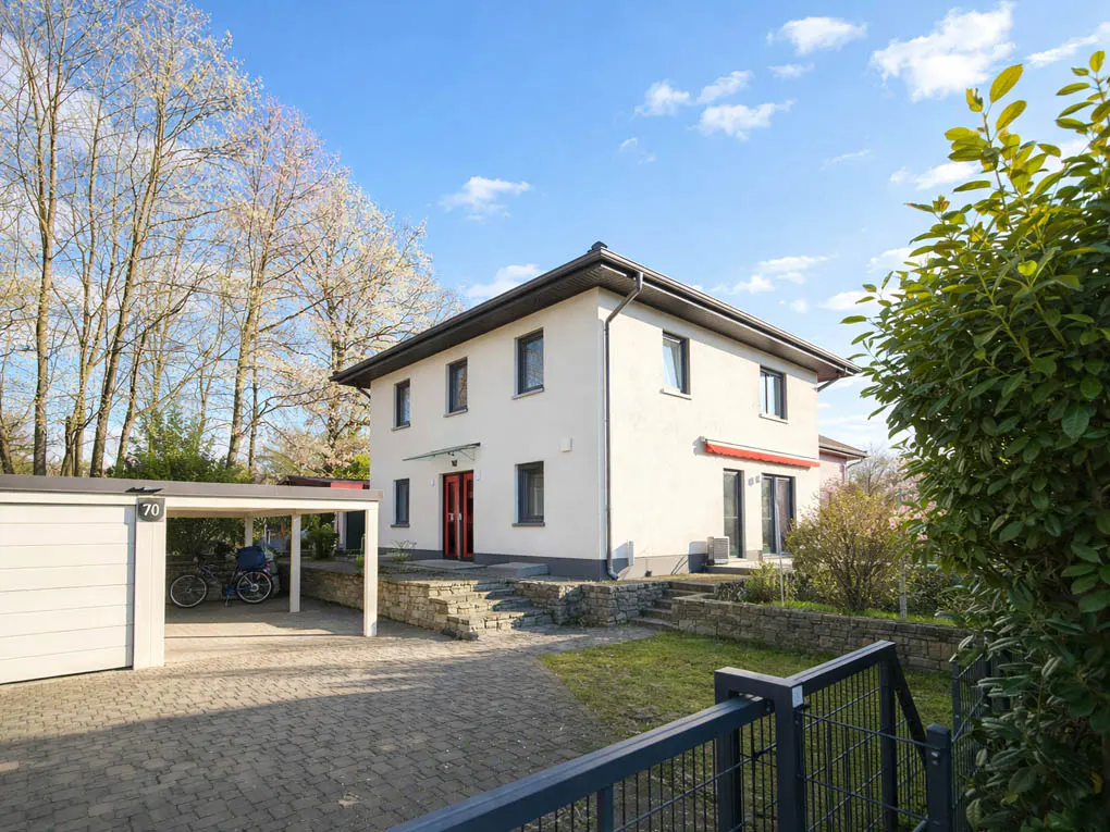 Two-story white house with a red door and a garage with bicycles under a carport. Blue sky.