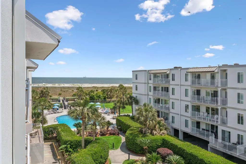 Condo view of pool, beach, and ocean. White building with balconies, palm trees, and blue sky.