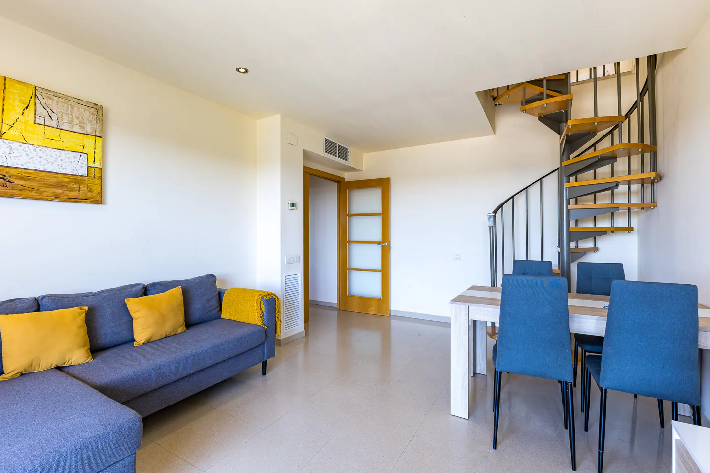 Modern living room with a grey sectional sofa, yellow cushions, dining table with blue chairs, and a metal spiral staircase.