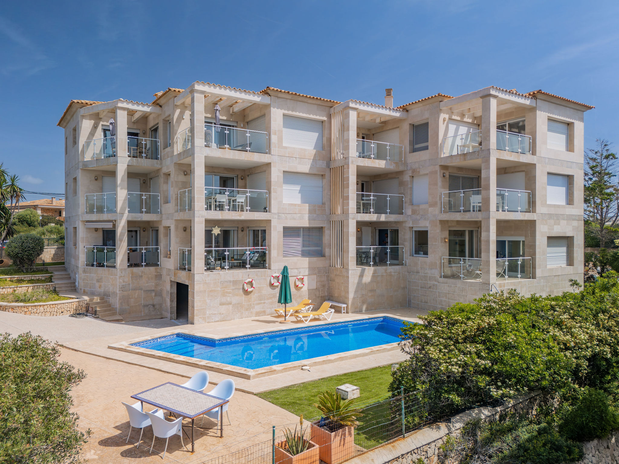 Exterior view of a light stone apartment building with balconies, a blue pool, and outdoor furniture.