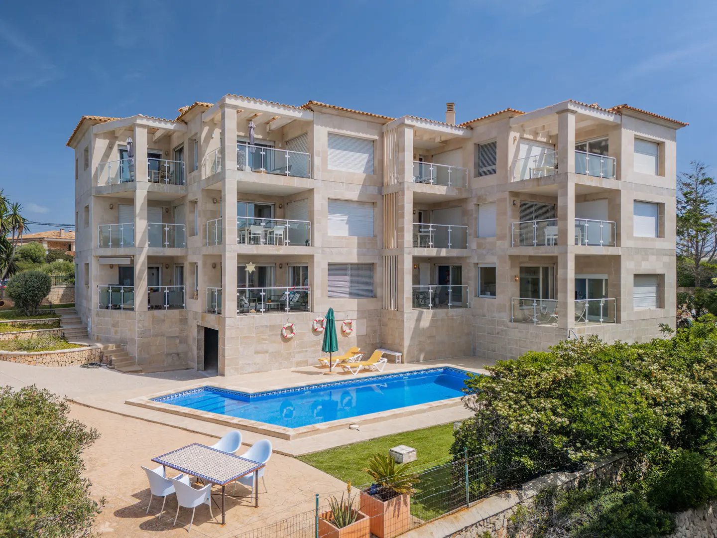 Exterior view of a light stone apartment building with balconies, a blue pool, and outdoor furniture.