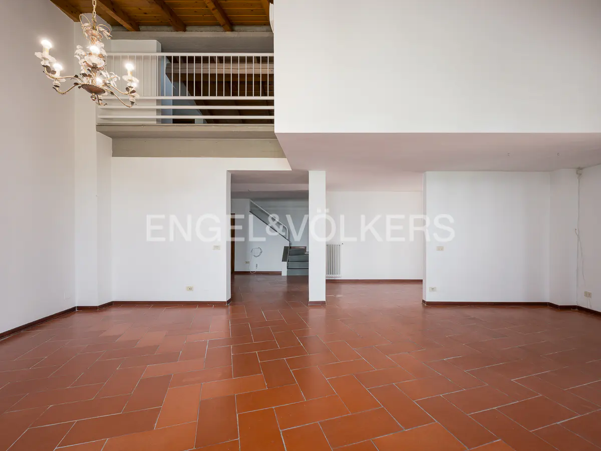 Empty room with white walls, terracotta tile floor, and a chandelier. A staircase is visible in the background.