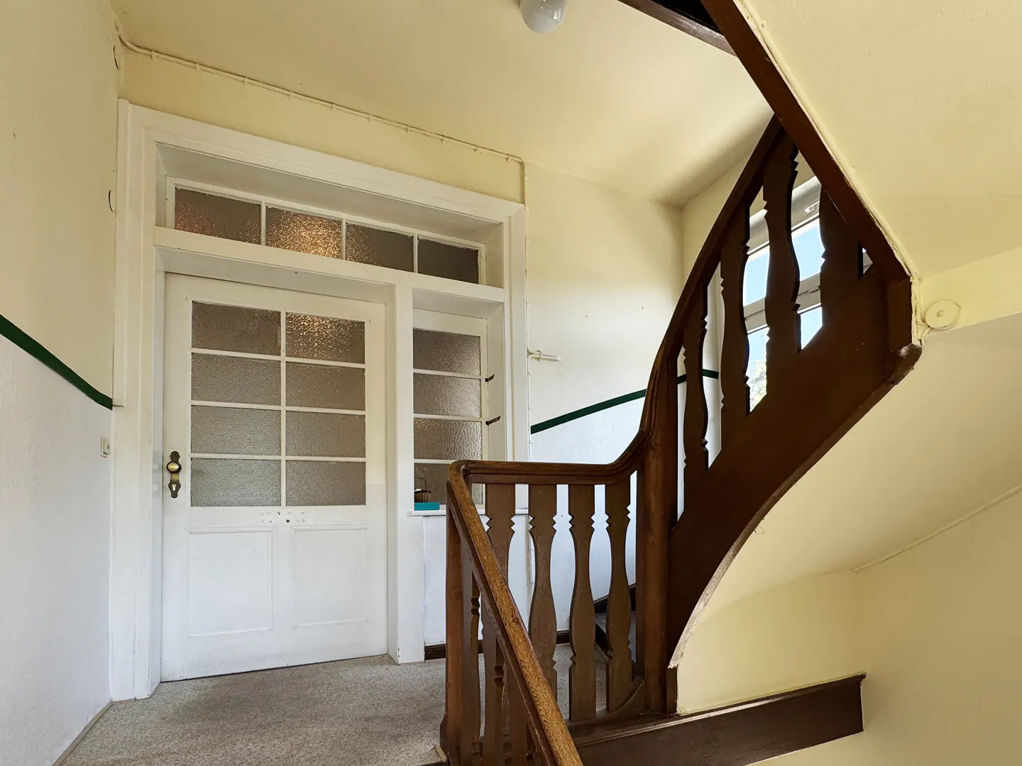 Interior view of a foyer with a white door with glass panels and a dark wood staircase with balusters. The walls are cream-colored with a green stripe.