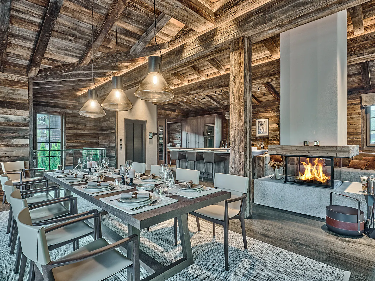 Rustic dining room with wood walls, beams, and a long table set for a meal. A modern fireplace glows in the background.