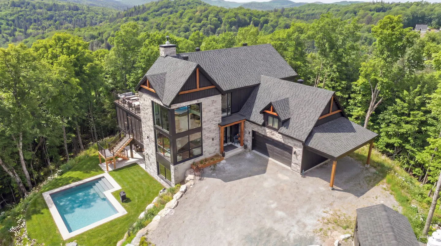 Aerial view of a modern stone house with a dark gray roof, surrounded by green trees, and a blue pool.