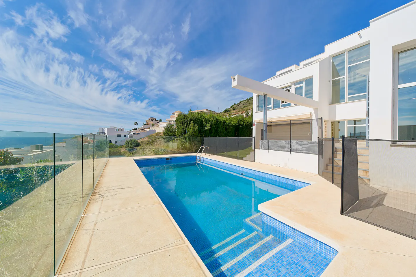 A modern white house with a blue tiled pool and glass fence under a blue sky with wispy clouds.