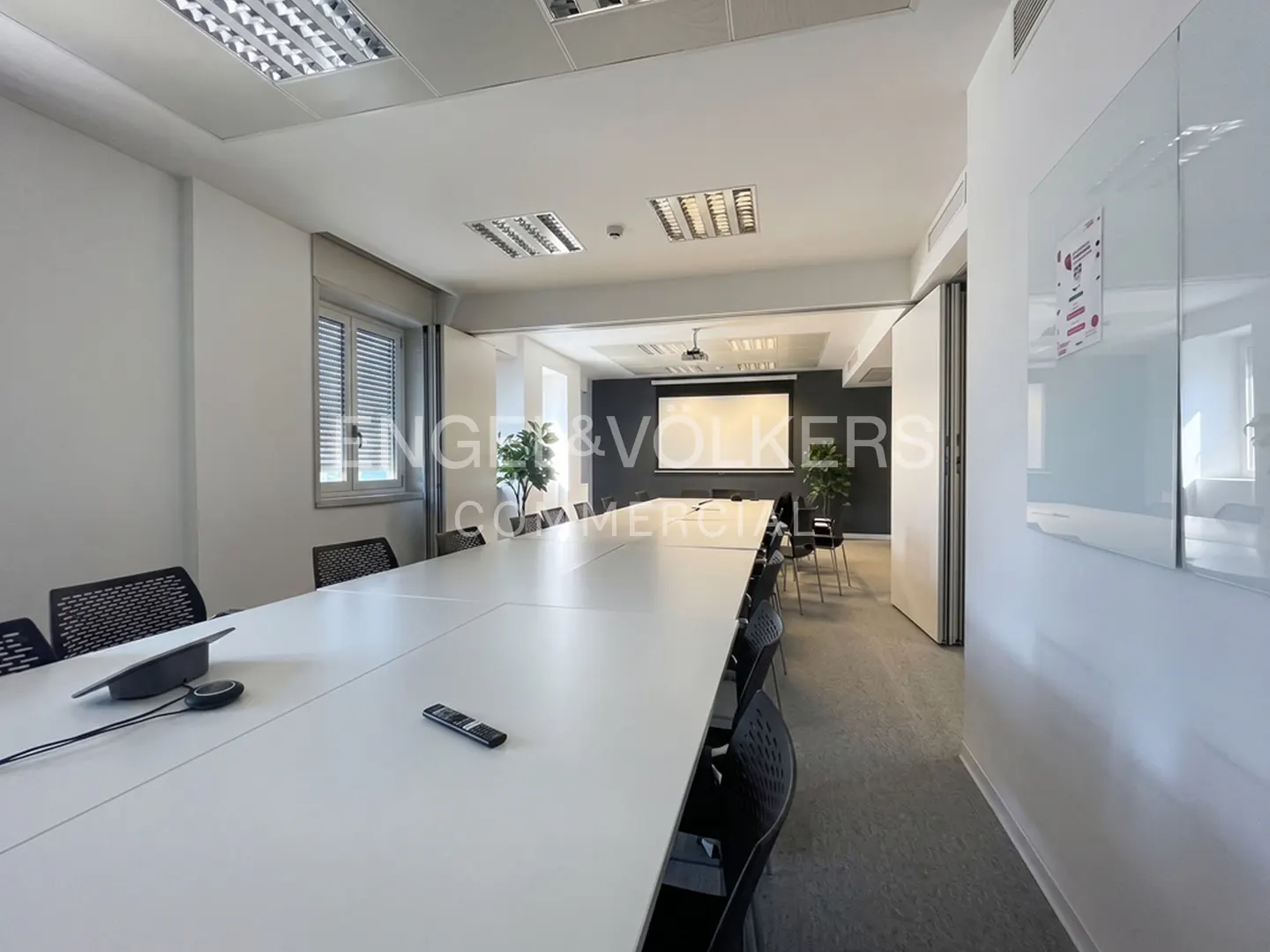Bright conference room with a long white table, black chairs, and a projector screen at the end.