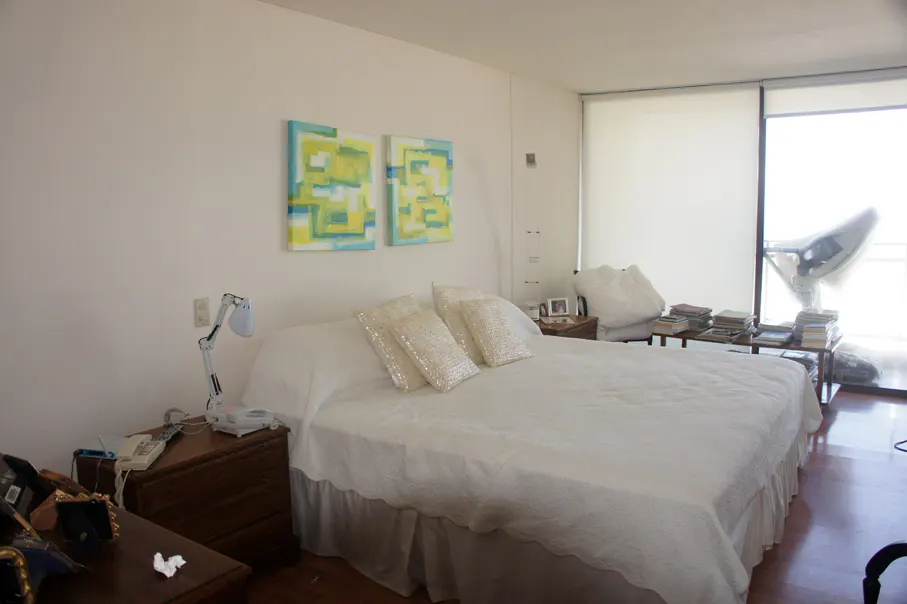 Bedroom with white walls, a large bed with white linens, and two abstract paintings in blue and yellow above the bed. A table with books is near a bright window.