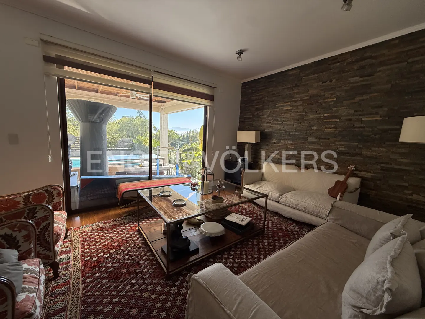 Living room with a large window view of a pool. A stone wall, sofas, and a glass coffee table are visible.