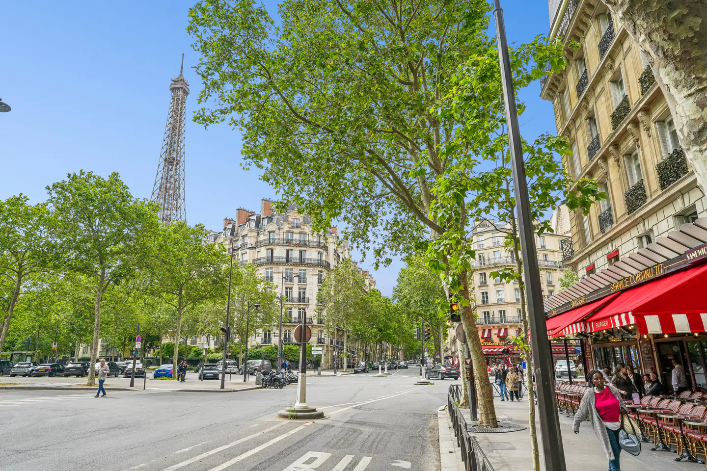 Paris street scene with the Eiffel Tower in the background. Buildings, trees, and people are visible. A red awning covers a restaurant.