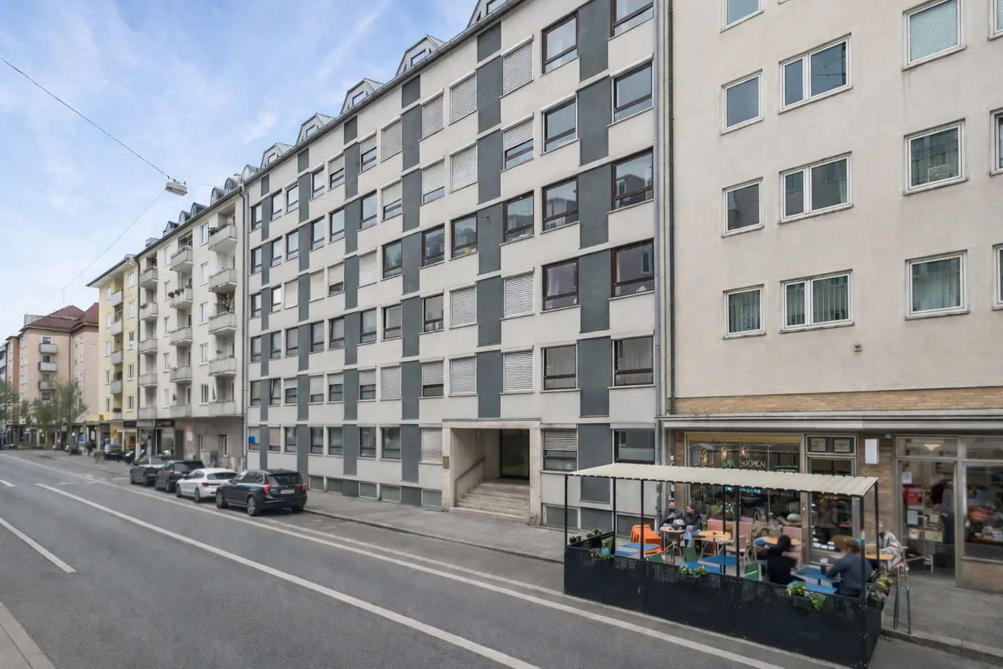 Street view of a multi-story apartment building with a cafe in front. Cars are parked along the street.