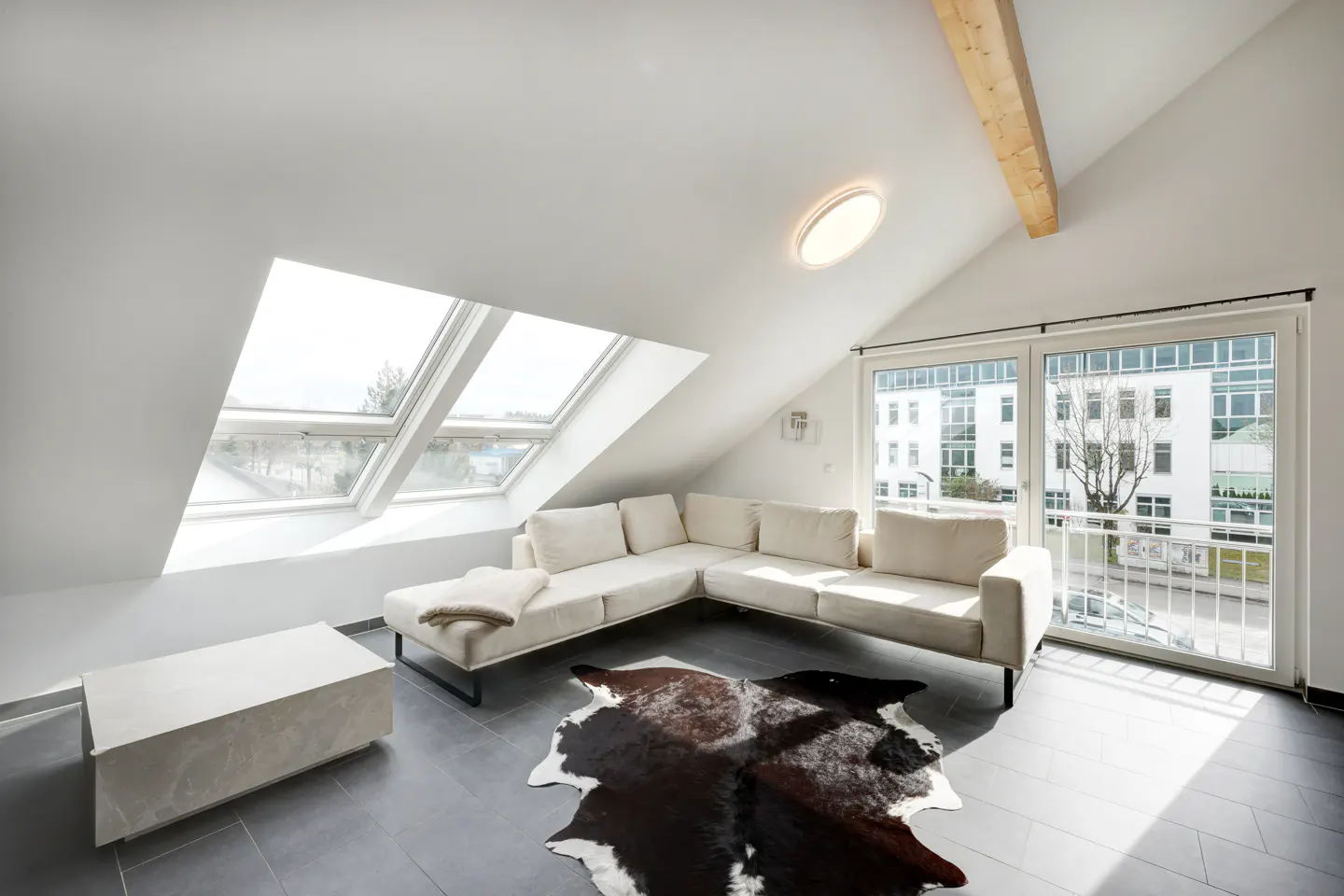 Bright attic living room with white walls, skylights, and a sliding glass door. A cream sectional sofa sits on a cowhide rug.