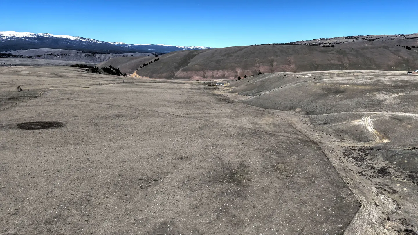 Vast, dry, tan field with hills and mountains in the background under a clear blue sky. Snow-capped mountains are visible in the distance.