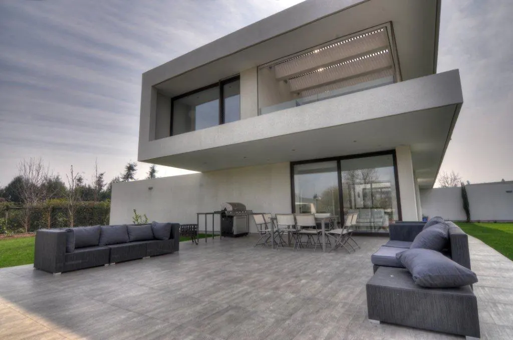 Modern two-story house with a gray patio featuring outdoor seating, a grill, and a dining table.