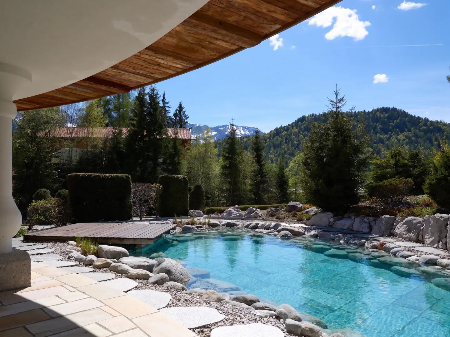 A natural pool with turquoise water, surrounded by rocks and greenery, with mountains in the background.