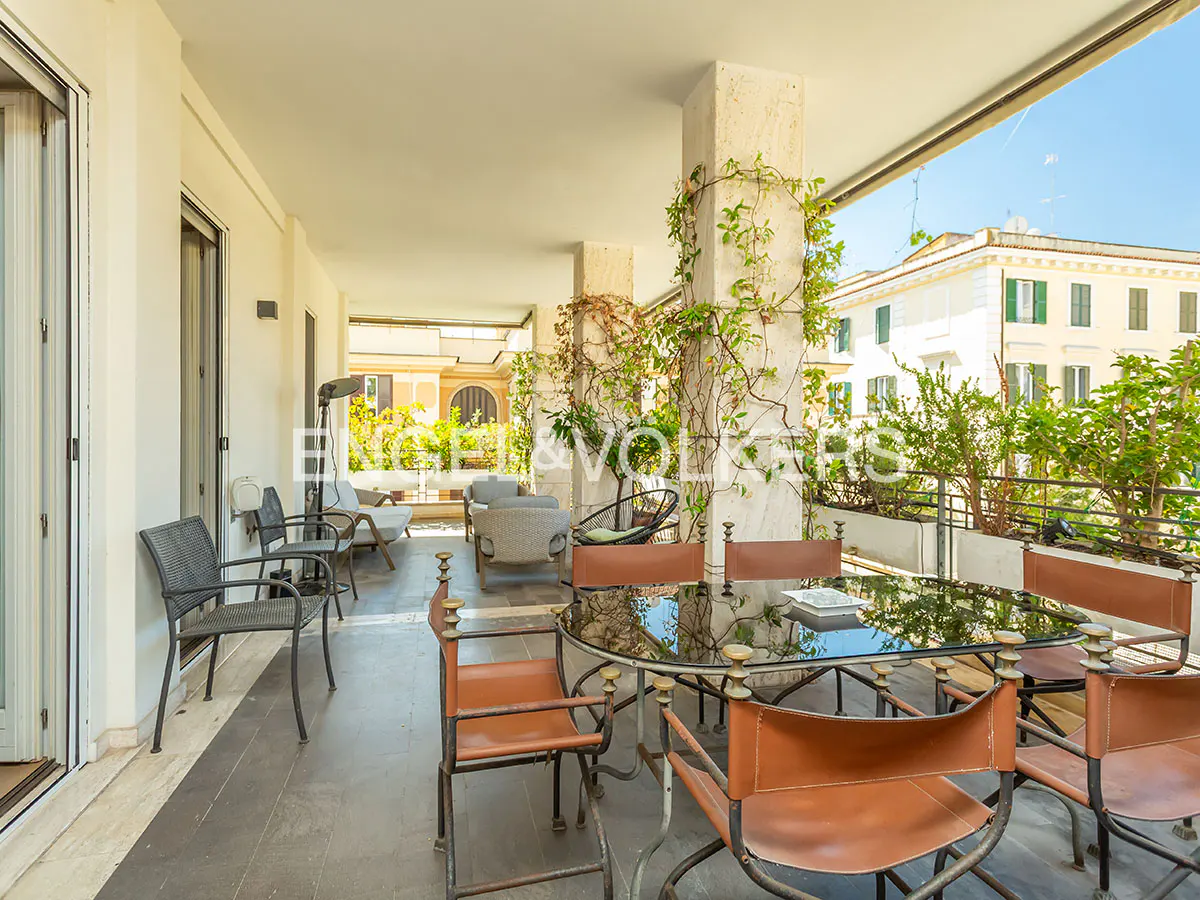 Spacious covered balcony with a glass dining table, tan leather chairs, and potted plants overlooking city buildings.