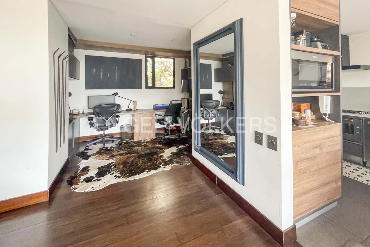 A modern office space with wooden floors, desks, and a cowhide rug. A large mirror reflects the room, next to a wooden cabinet with a microwave.