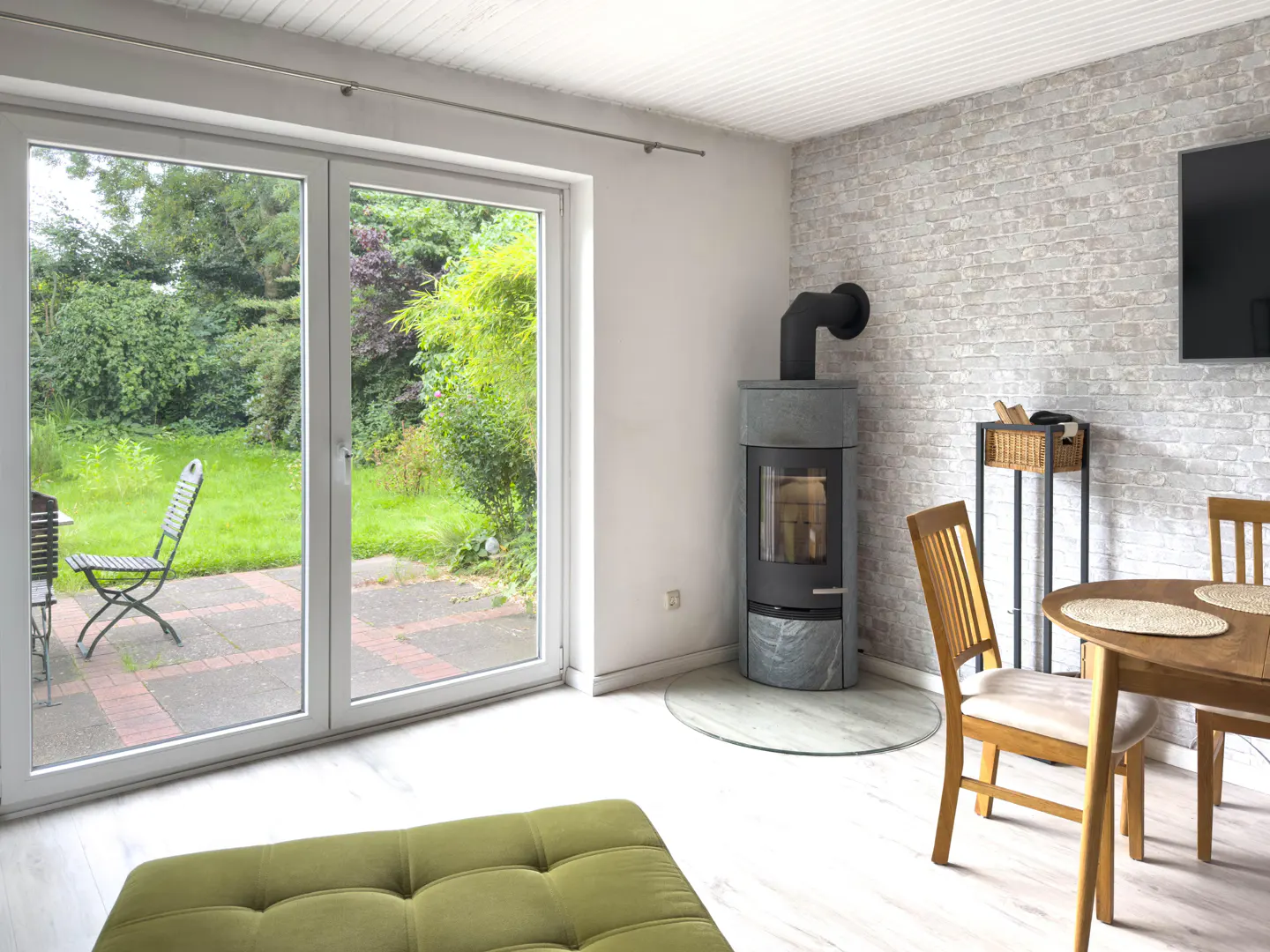 Bright living room with white walls, wood floors, and a stone fireplace. Large sliding glass doors open to a green garden with patio furniture.