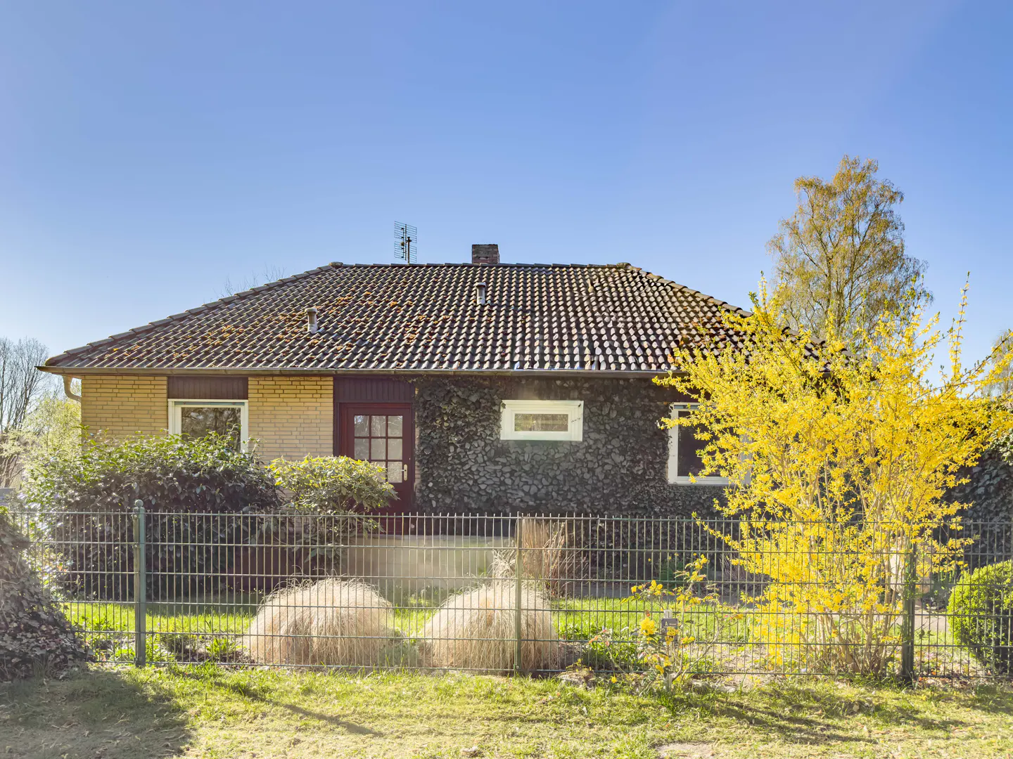 A single-story brick house with a brown tile roof, a red door, and a green metal fence in front. A yellow flowering bush is on the right.