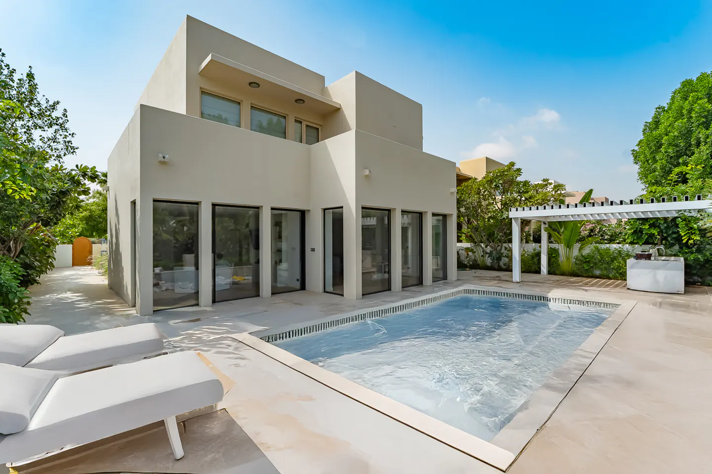 Backyard view of a modern, two-story beige house with a pool, lounge chairs, and a white pergola.