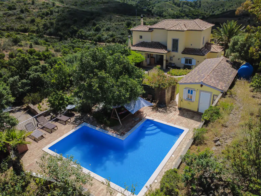 Aerial view of a yellow two-story house with a pool, surrounded by trees and greenery.