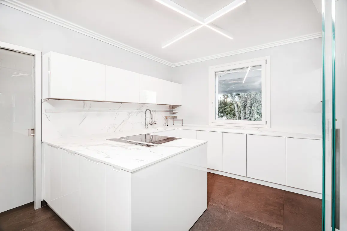Bright, modern kitchen with white cabinets, marble countertops, and a central island. A window offers a view of trees.