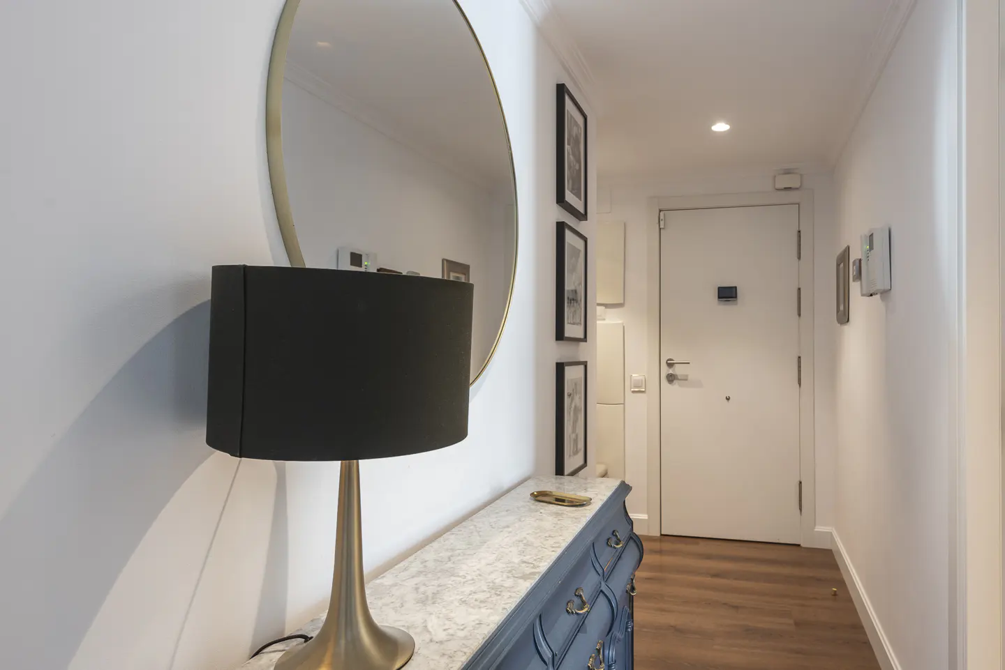 Hallway with white walls, wood floor, blue dresser, gold lamp, round mirror, and white door.
