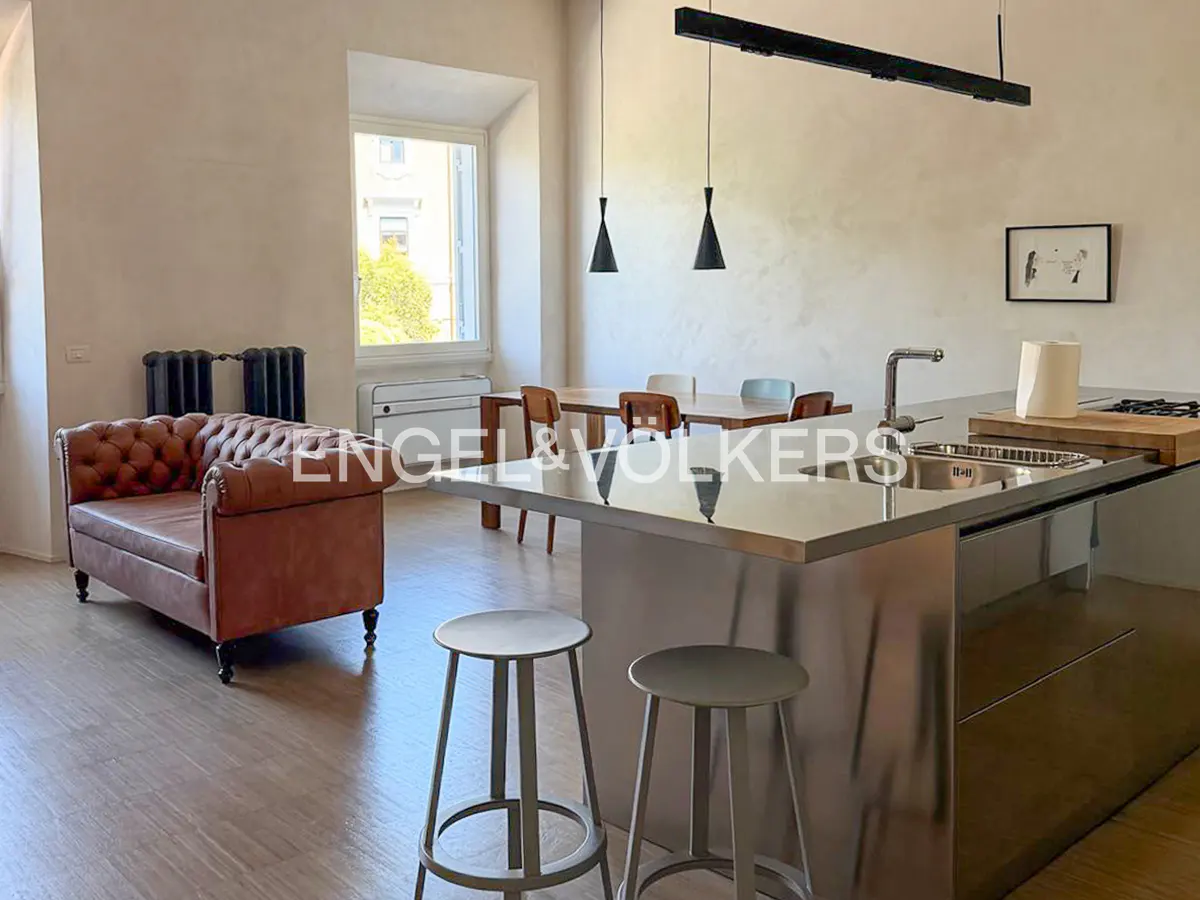 A modern apartment interior with a brown leather sofa, stainless steel kitchen island with stools, and a dining table.