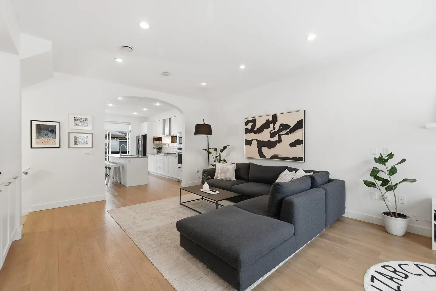 Bright, open-concept living room with hardwood floors, a gray sectional sofa, and a view into the white kitchen. Abstract art hangs on the wall.